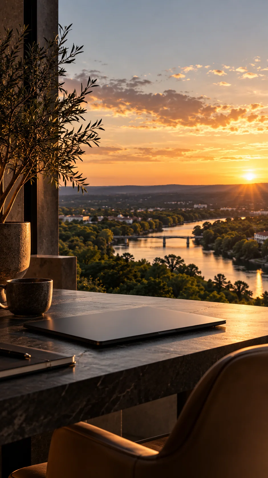 A closed laptop and coffee cup on a stone desk above a river at sunset